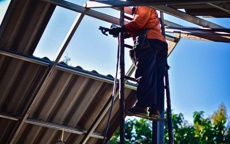 Trabalhador em uma escada realizando manutenção no telhado, usando equipamentos de proteção e ferramentas, com céu azul ao fundo.